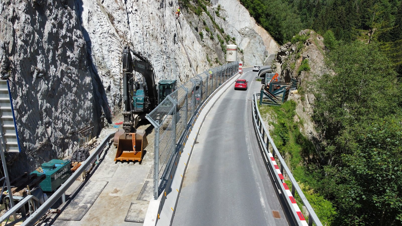 Protective barrier between a steep rock wall and a road, vehicles on the road and an excavator on the left